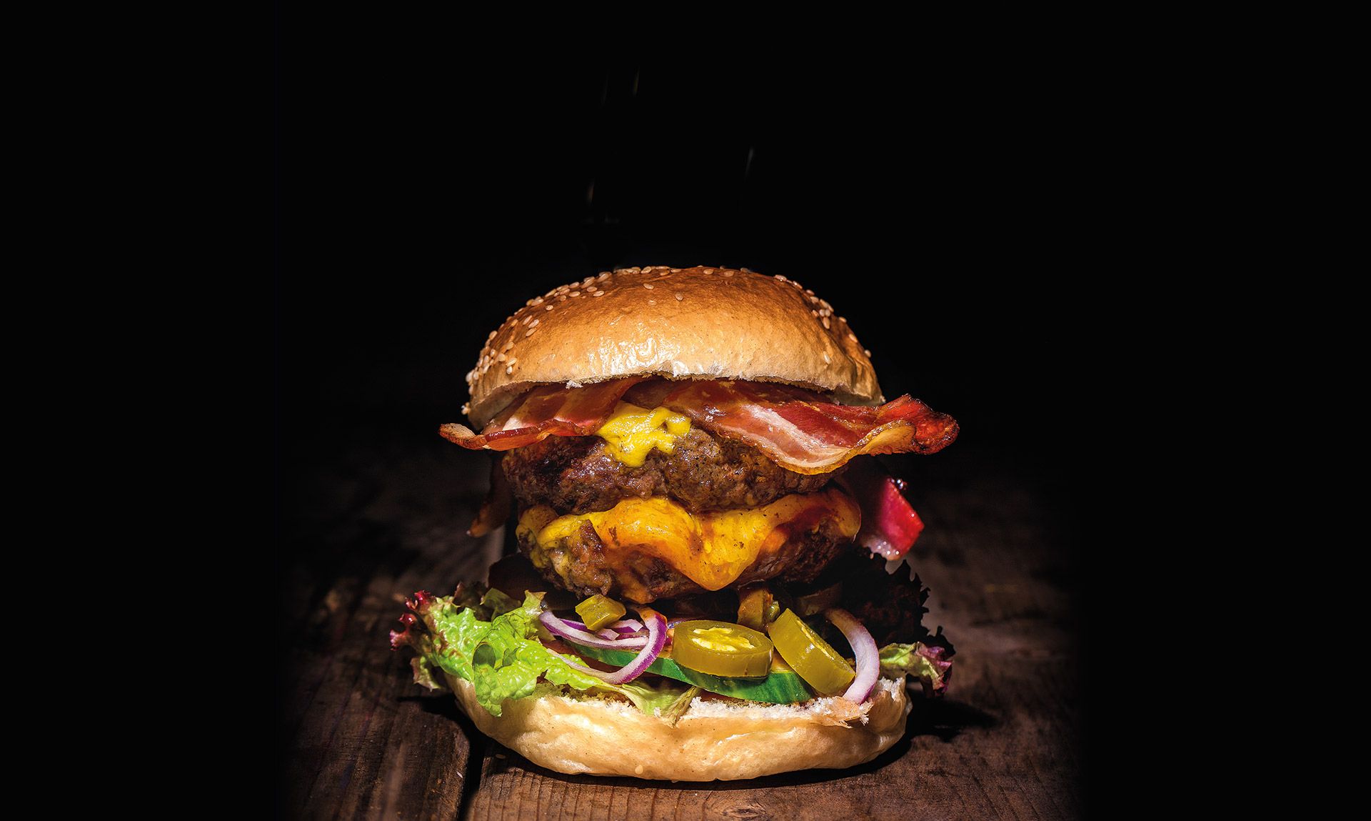 Hoher Burger mit Cheddar, Bacon, Jalapeños und marinierten Zwiebeln auf einem Holzbrett